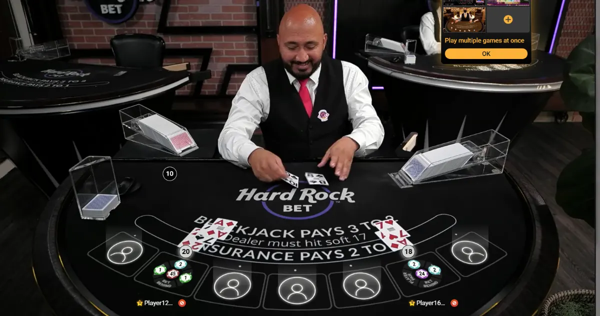 Live blackjack scene with a male dealer dealing cards at a black “Hard Rock Bet” table, showing player hands, chips and betting spots on the felt, with card shoes on both sides and a small on-screen “Play multiple games at once” prompt.