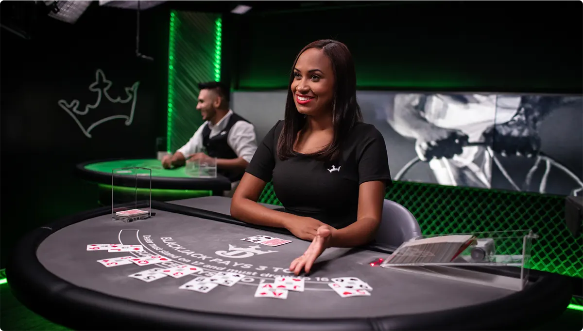 Smiling live blackjack dealer sits at a grey felt table with scattered playing cards and a shoe, set in a dark studio with green neon lighting and another table in the background.